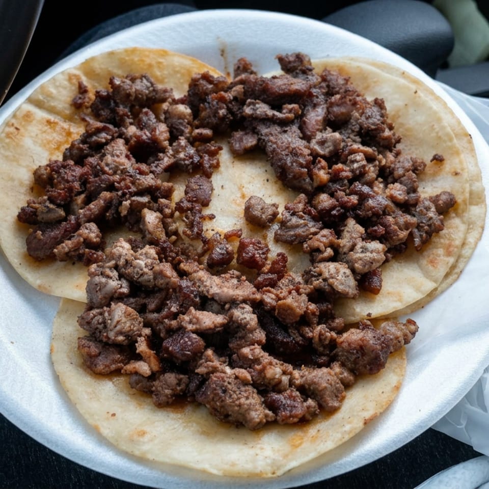 Three corn tortillas topped with browned chopped meat on a disposable plate inside a car.