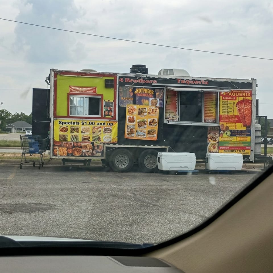 Black-and-green Mexican taqueria food truck parked beside a commercial building, with menu signs, coolers, and shopping carts outside.